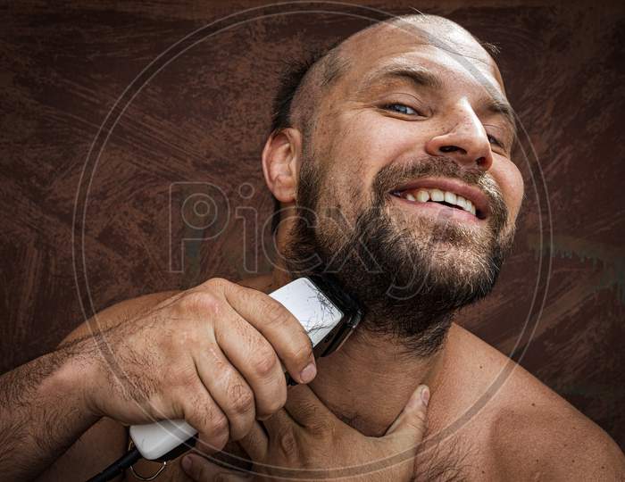 Caucasian Man Trying To Shave With An Electric Razor. A Brutal Bald Man Holds A Razor In His Hand And Shaves Stubble On A Metal Background
