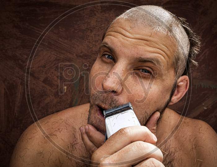Caucasian Man Trying To Shave With An Electric Razor. A Brutal Bald Man Holds A Razor In His Hand And Shaves Stubble On A Metal Background