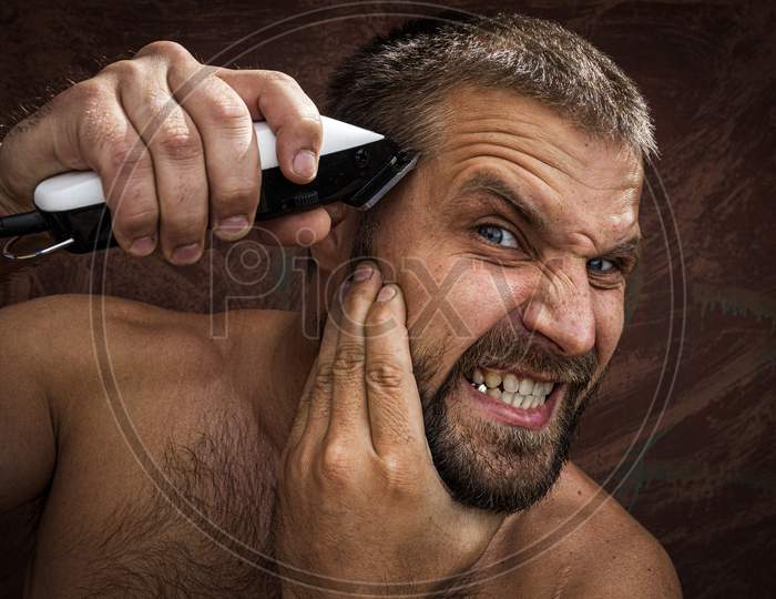 Close-Up Portrait Of Handsome Shirtless Man Shaving His Head With An Electric Razor And Gritting His Teeth, Against Brutal Background. Concept Of Male Home Care Without Beauty Salons