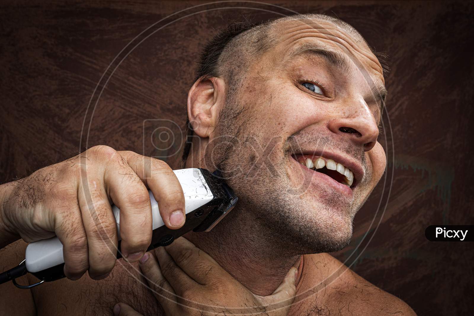 Adult Cheerful Man With A Beard Shaves His Beard At Home. The Guy Looks In The Bathroom Mirror And Uses The Electric Trimmer. Front View