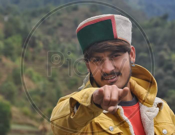 A good looking young guy showing rock gesture with wearing yellow jacket and himachali topi (Himachali traditional cap), A Indian young men standing outdoor with beautiful mountains view in background