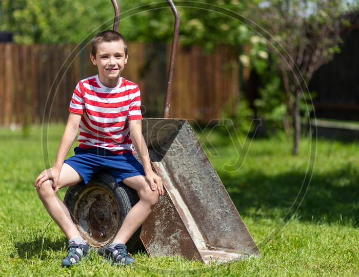 A Little Boy Sitting On The Wheelbarrow   In A Sunny  Day In The Garden .Boy Assistant In The Garden