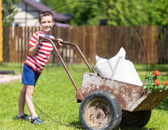 Cute Toddler Boy Playing With Big Old Wheelbarrow At Backyard In Garden Outdoors.Young Smile Boy Pushes A Wheelbarrow Around A Yard.