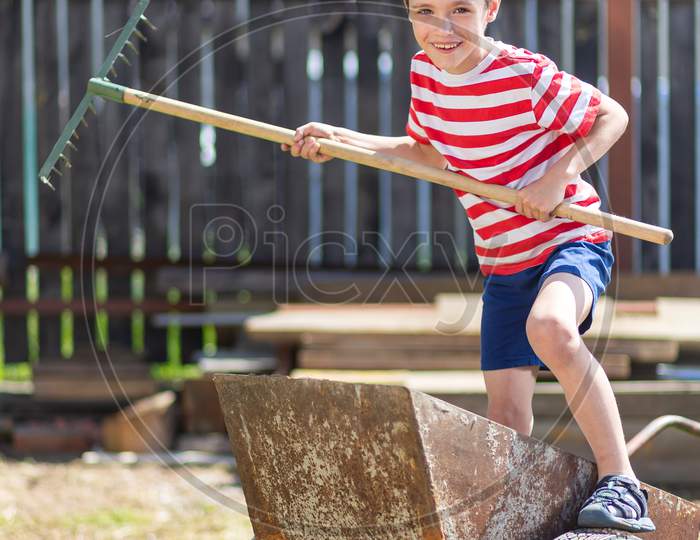 A Little Cheerful Boy Stands On A Garden Wheelbarrow And Holds A Rake In His Hand Like A Paddle In The Garden Of A Country House. The Boy Introduces Himself As The Captain In The Garden