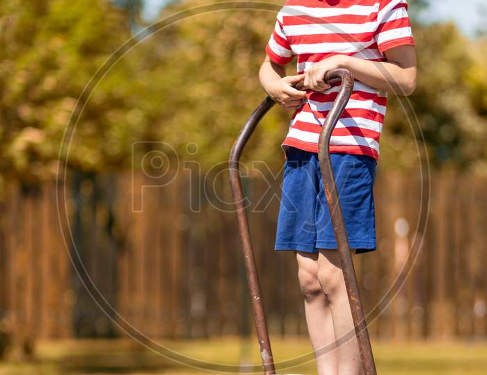A Little Cheerful Boy Stands On A Garden Wheelbarrow And In The Garden Of A Country House. Little Helper Boy Ready To Work