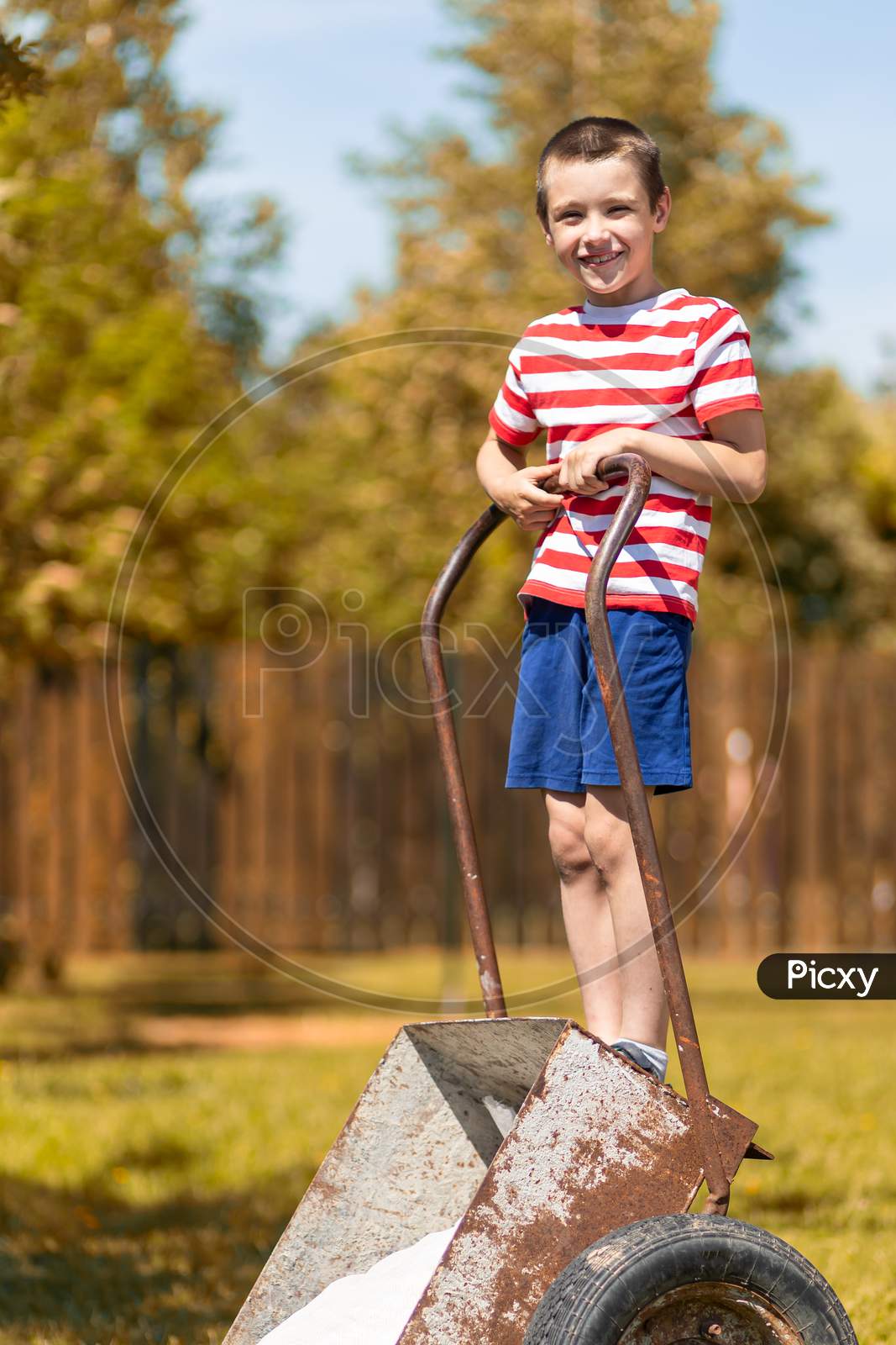 A Little Cheerful Boy Stands On A Garden Wheelbarrow And In The Garden Of A Country House. Little Helper Boy Ready To Work