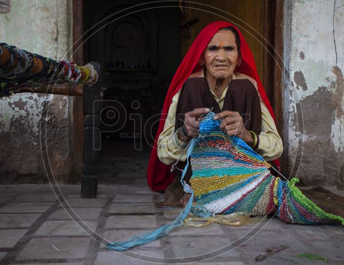 An aged woman wearing traditional dress, sitting in front of house and weaving