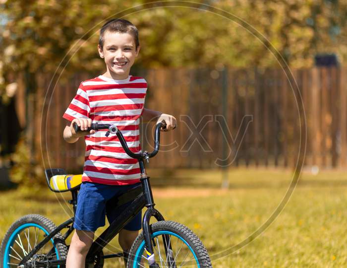 Happy Cheerful Boy In A Striped T-Shirt Is Doing Well, Looking At The Camera And Sitting On A Bike Against The Background Of The Garden. Active Outdoor Games For Children In Summer.
