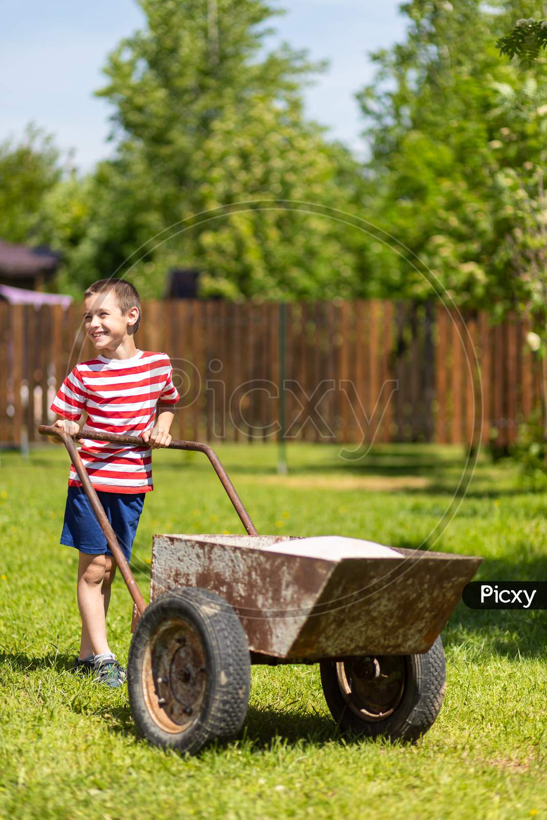A Little Boy Drives A Wheelbarrow With Bags On A Sunny Day In The Garden. Boy Assistant In The Garden