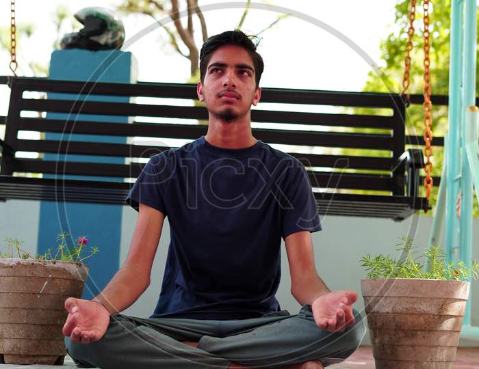 A Boy Is Meditating At Home In India. Indian Young Boy In A Black Traditional Shirt Sitting In A Yoga Position.