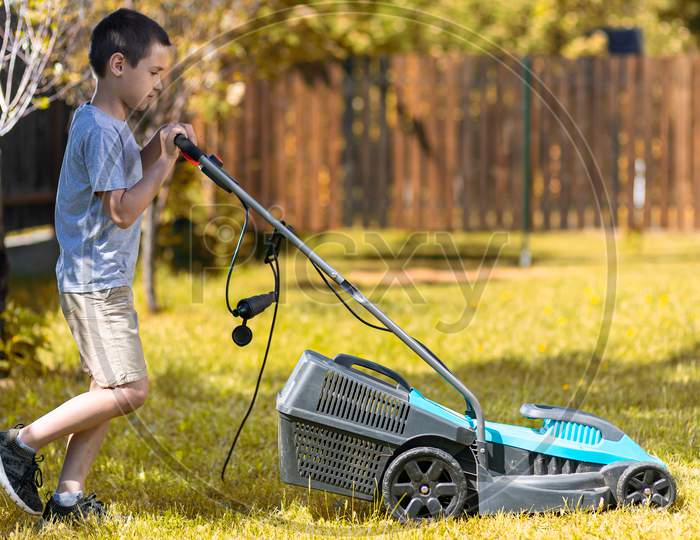 A Boy  With An Electric Lawn Mower Mowing The Lawn.Beauty Boy  Pruning And Landscaping A Garden, Mowing Grass, Lawn, Paths.