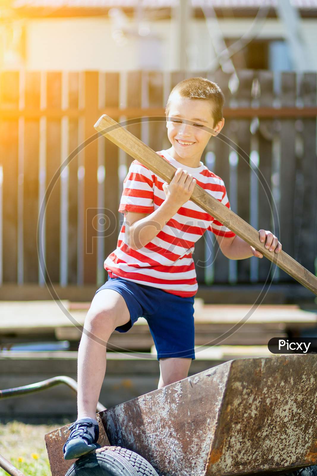 A Little Cheerful Boy Stands On A Garden Wheelbarrow And Holds A Shovel In His Hand Like A Paddle In The Garden Of A Country House. The Boy Introduces Himself As The Captain In The Garden