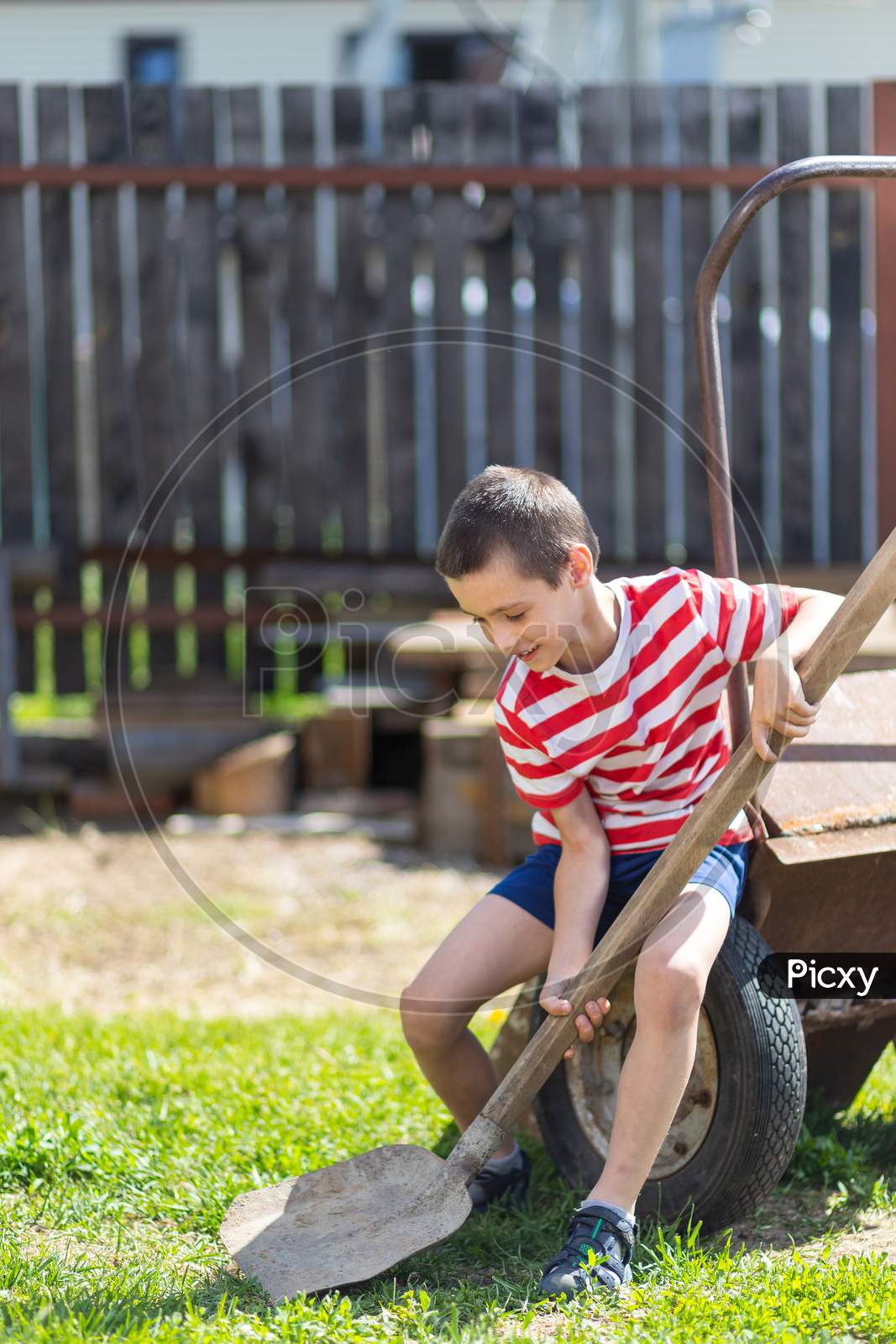 A Little Cheerful Boy Sits On A Garden Wheelbarrow And Holds A Shovel In His Hand In The Garden Of A Country House. Little Boy Helper Ready To Dig