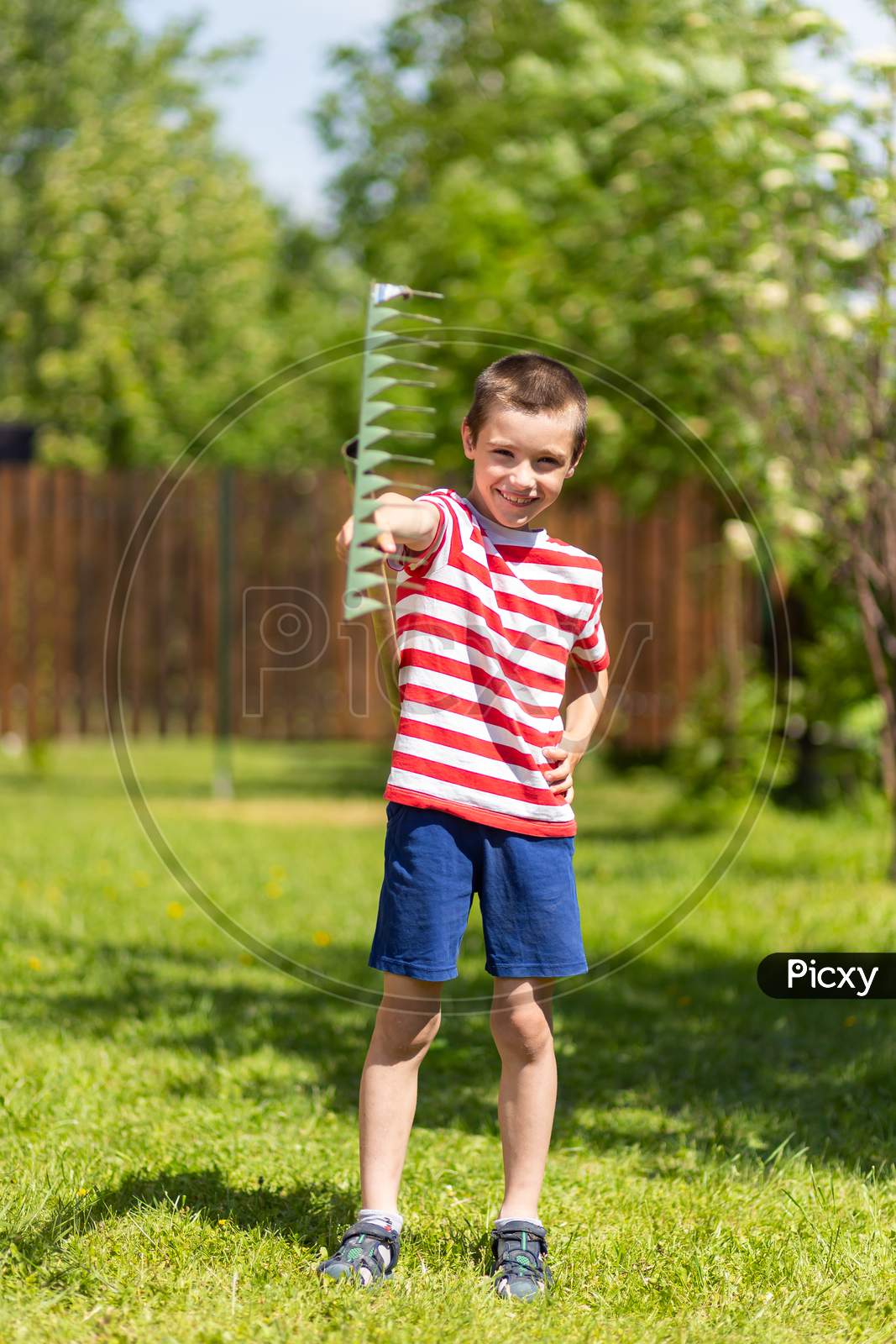 A Little Cheerful Boy Stands And Holds A Rake In His Hand, Ready To Work In The Garden Of A Country House.