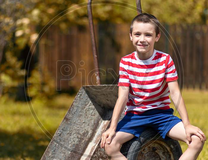 A Little Cheerful Boy Sits On A Garden Wheelbarrow In The Garden Of A Country House. Little Boy Helper Ready To Dig