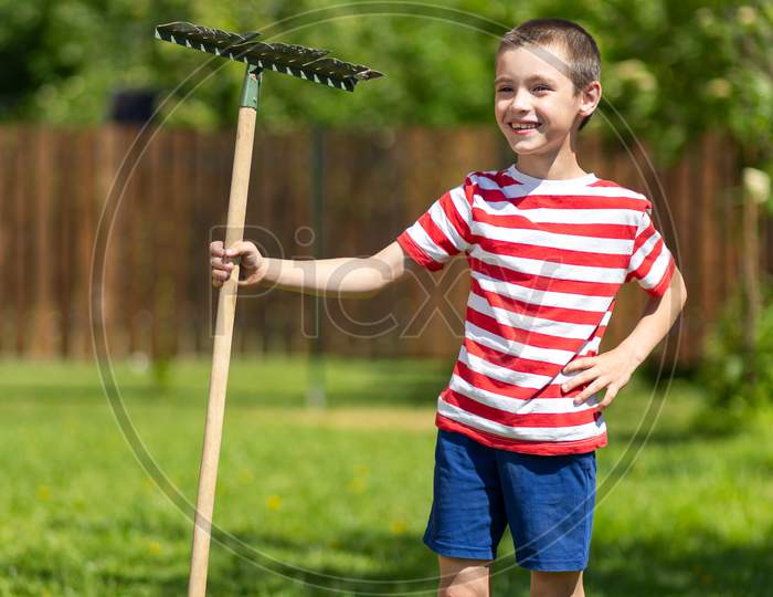 A Little Cheerful Boy Stands And Holds A Rake In His Hand, Ready To Work In The Garden Of A Country House.
