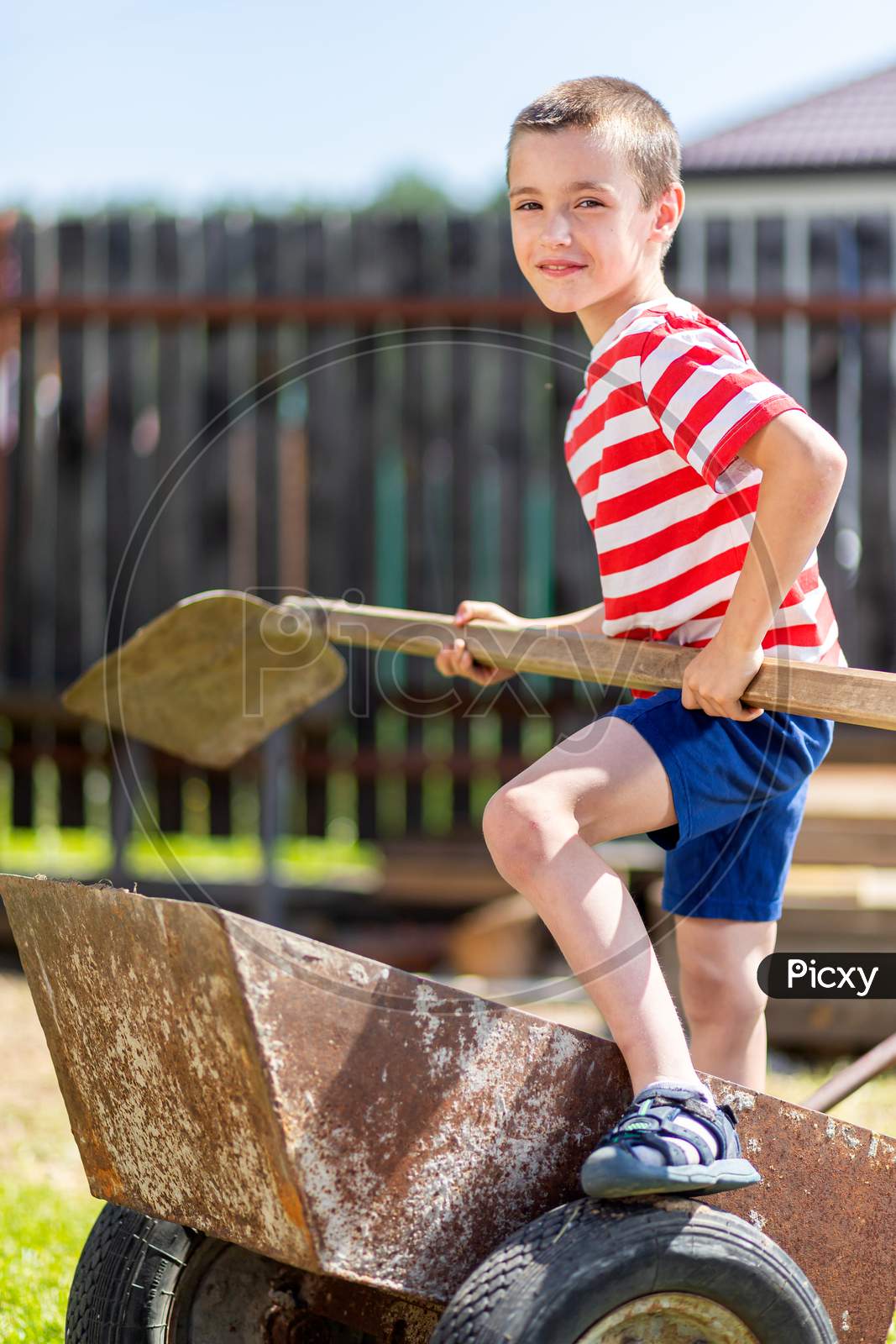 A Little Cheerful Boy Sits On A Garden Wheelbarrow And Holds A Shovel In His Hand In The Garden Of A Country House. Little Boy Helper Ready To Dig