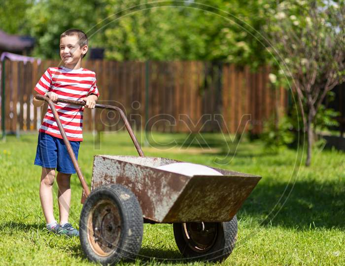 Young Smile Boy Pushes A Wheelbarrow Around A Yard.Boy Helper In T-Short And Shorts Having Fun Pushing Barrow And Gardening At Countryside