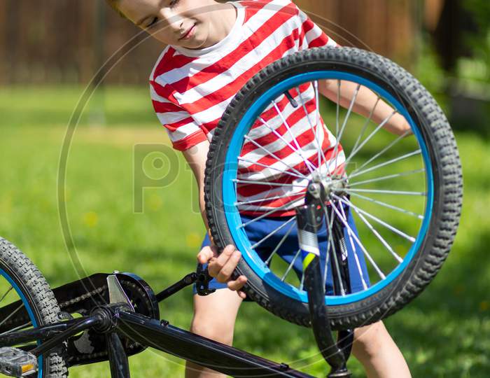 Happy Cheerful Boy In A Striped T-Shirt Feels Good, Looks At The Wheel And Tries To Repair The Bike Against The Background Of The Garden. Active Outdoor Games For Children In Summer.