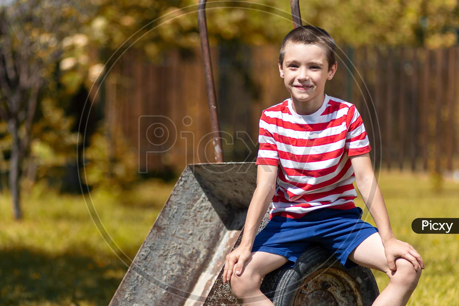 A Little Cheerful Boy Sits On A Garden Wheelbarrow In The Garden Of A Country House. Little Boy Helper Ready To Dig