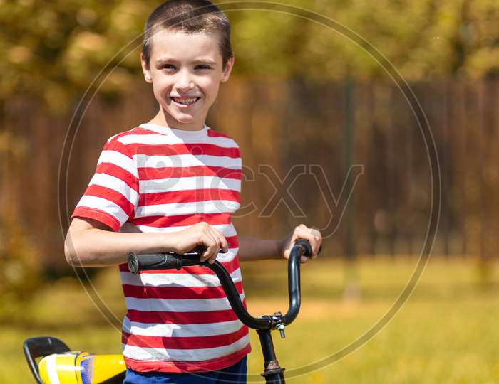 Happy Cheerful Boy In A Striped T-Shirt Is Doing Well, Looking At The Camera And Sitting On A Bike Against The Background Of The Garden. Active Outdoor Games For Children In Summer.