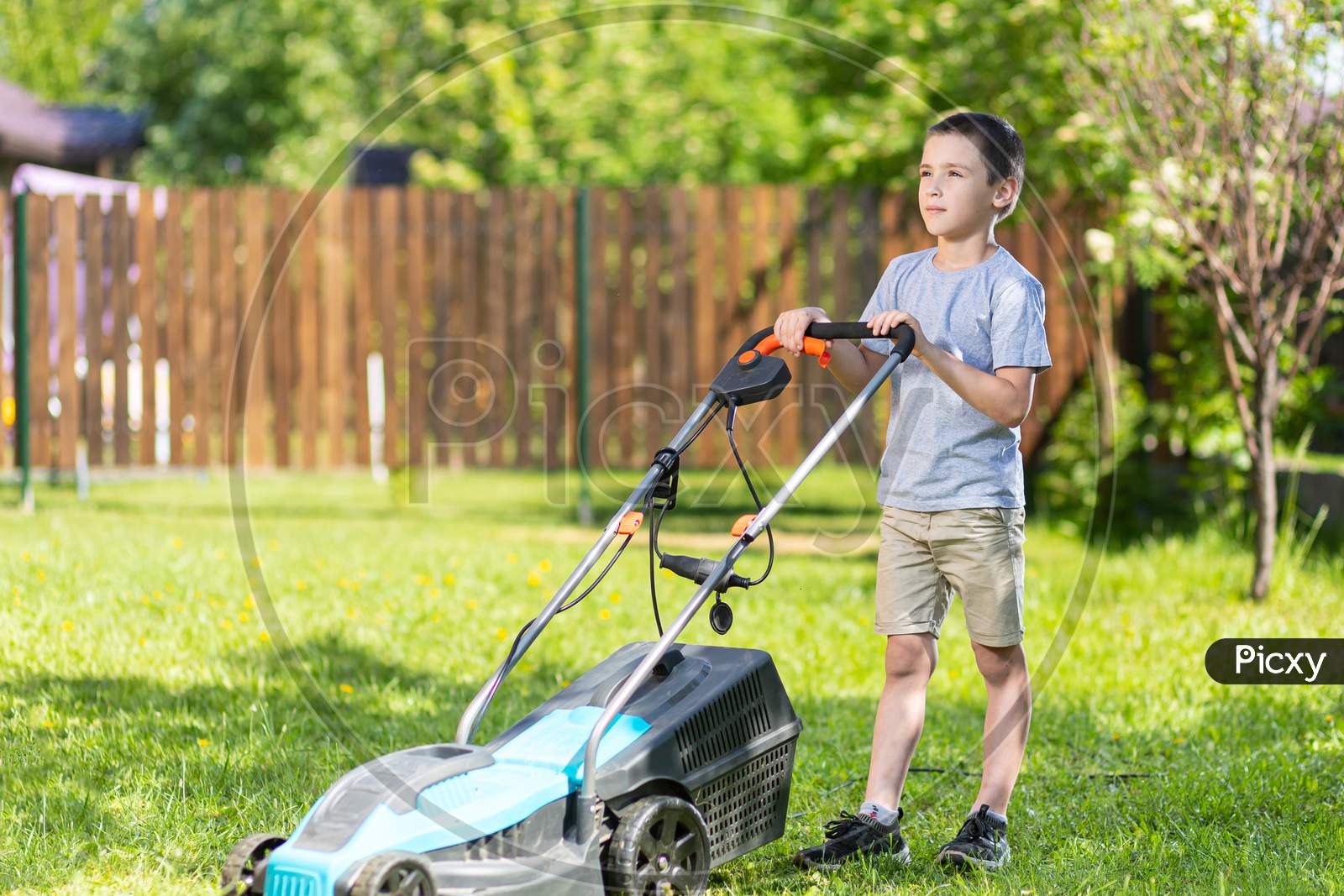 Boy Worker On The Garden Working On Mowing The Lawn With The Help Of A Modern Lawn Mower Near A Country House
