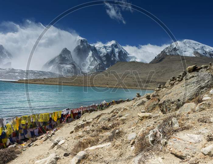 A Beautiful Landscape Of Gurudongmar Lake With Rocks In Forte Ground And Mountain With Blue Sky In Background. Gurudongmar Lake One Of The Highest Lake Of The World And India Located At An Altitude Of 17,800 Ft, In The Indian State Of Sikkim