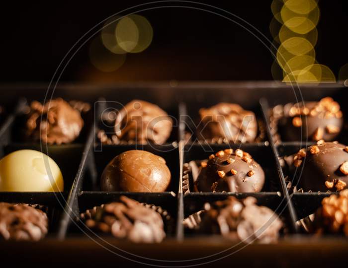 Delicious Chocolate Candies Or Truffle Balls In Box On Table Against Light Bokeh. Assorted Chocolate Pralines.An Assortment Of Fine Chocolates In White, Dark, And Milk Chocolate With Nuts.