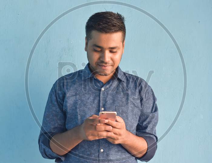 Portrait of young Indian guy holding smartphone and smiling looking at his smartphone. Isolated in a light blue background.
