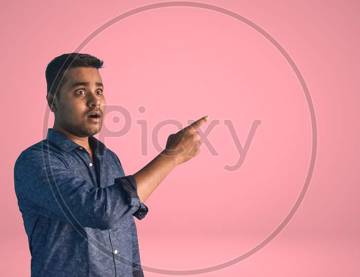 A young Indian man in shock Pointing his index finger aside in semi-casual clothes posing isolated on a light pink background. Surprised young man. Brand endorsement concept.