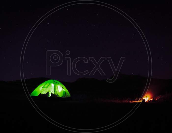 Illuminated Green Tent Standing By Camp Fire In Nature At Night With Starry Night Background.