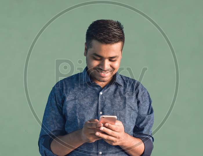 Portrait of young Indian guy holding smartphone and smiling looking at his smartphone Isolated in a blue background. Happy businessman concept.