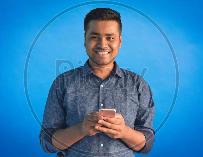 Portrait of young Indian guy holding smartphone and smiling looking at camera Isolated in a blue background. Indian businessman and finance concept.