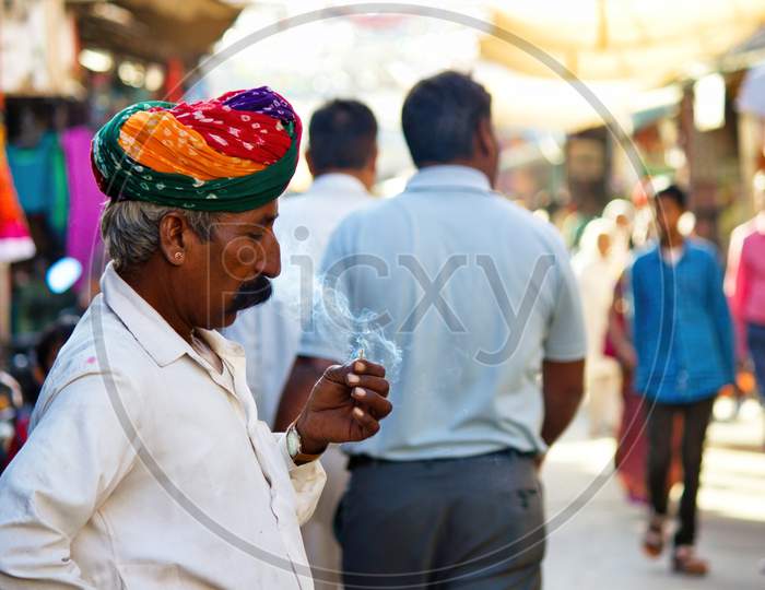 Pushkar, India - November 10, 2016: An Old Rajasthani Man In Traditional Ethnic Wear Such As Colorful Turban And Typical White Shirt Smoking Cigarette Or Mini-Cigar Filled With Tobacco
