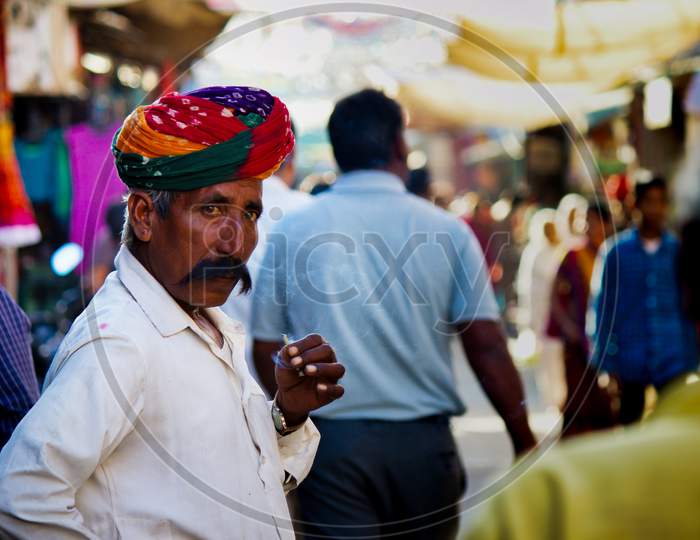 Pushkar, India - November 10, 2016: An Old Rajasthani Man In Traditional Ethnic Wear Such As Colorful Turban And Typical White Shirt Smoking Cigarette Or Mini-Cigar Filled With Tobacco