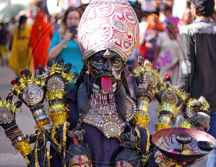 Pushkar, India - November 10, 2016: A Young Little Girl Dressed Up Or Disguised As Indian Goddess Of Destruction Named Maa Kali With Crown And Multiple Hands As Street Performer During Pushkar Fair