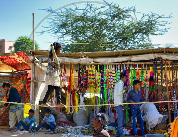 Pushkar, India - November 10, 2016: A Little Gypsy Indian Girl Doing Show Of Walking Bare Foot On A Rope Performing Acrobatic Balancing Act While Holding Bamboo Stick In Her Hand During Pushkar Fair
