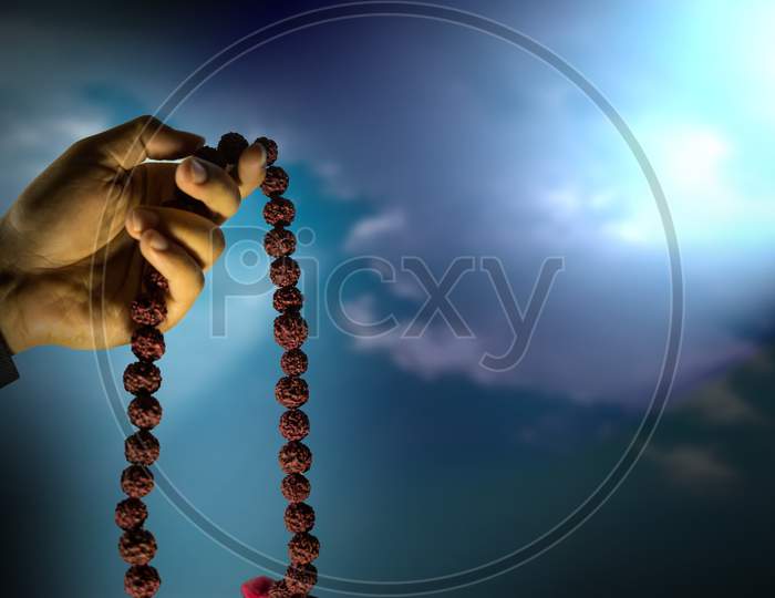 Prayer Beads In Hand Isolated On A Blue Sky With Sun Rays . Male Hand Holding Rosary, Praying To God, Religious Spirituality. Man Holding Rosary Beads And Meditating. Religious Concept