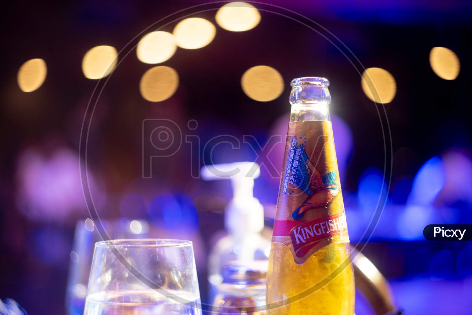 Famous Indian Beer Brand Kingfisher Bottle With Bubble Coming Out With Out Of Focus Background Lights With Bokeh Balls Showing It Placed In A Bar Club Pub Nightlife Spot In Bangalore, Mumbai With Alcohol Placed All Around