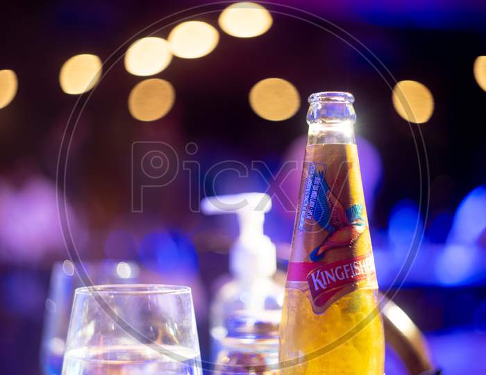 Famous Indian Beer Brand Kingfisher Bottle With Bubble Coming Out With Out Of Focus Background Lights With Bokeh Balls Showing It Placed In A Bar Club Pub Nightlife Spot In Bangalore, Mumbai With Alcohol Placed All Around