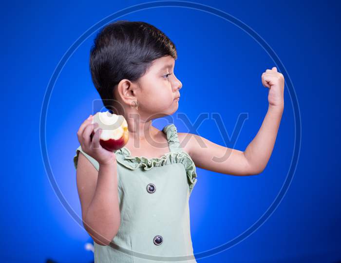Cute Little Girl Eating Apple And Showing Muscle Biceps On Blue Studio Background - Concept Showing Of Healthy Food Or Eating Fruits Makes Stronger.