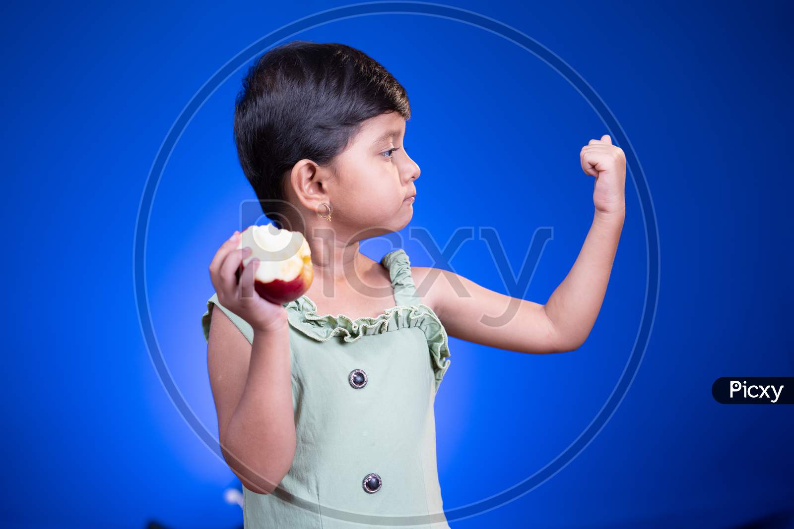 Cute Little Girl Eating Apple And Showing Muscle Biceps On Blue Studio Background - Concept Showing Of Healthy Food Or Eating Fruits Makes Stronger.