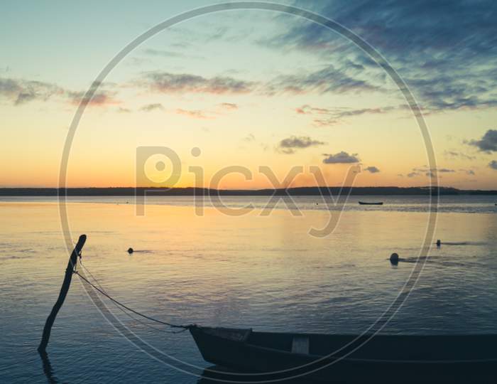 Small Boat Anchored On The Shore Of The Lake During A Day'S Sunset With Some Clouds. Calm Waters
