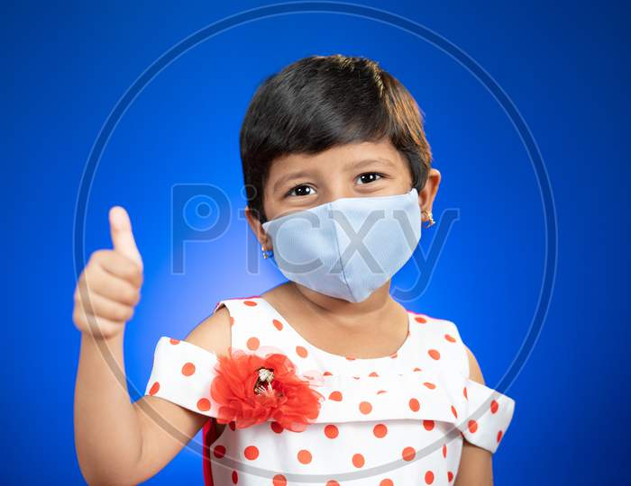 Little Girl Kid Adjusting Medical Face Mask And Showing Thumps Up Gesture - Concept Showing Of Coronavirus Covid-19 Saferty Measures By Wearing Mask As New Normal