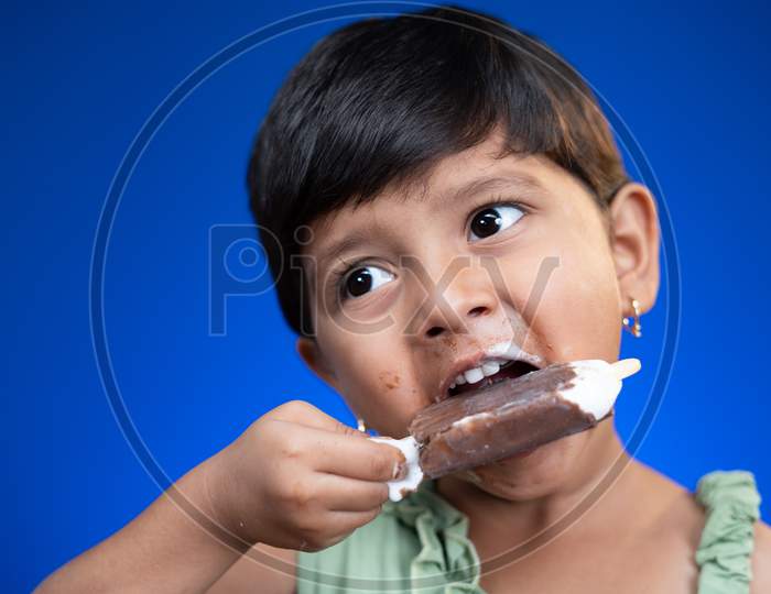 Cloe Up Head Shot Of Girl Kid Busy Eating Ice Cream On Blue Studio Background - Concept Of Unhealthy Food Consumption
