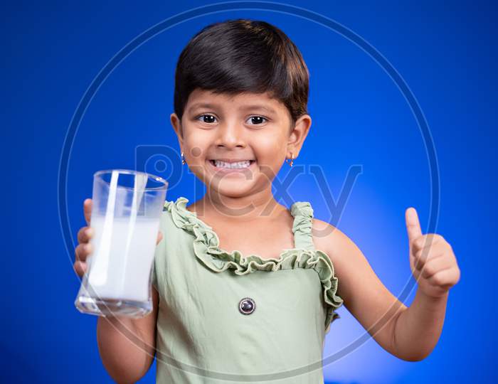 Little Girl Kid With Milk In Hand Showing Thumbs Up Sign On Blue Background - Concept Showing To Encouragement Or Recommending Milk For Children's Health