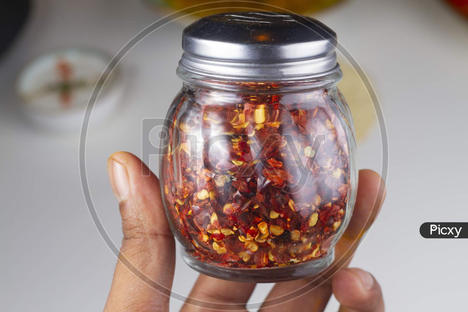 Chili Flakes Jar In Hand Isolated On Light Background.