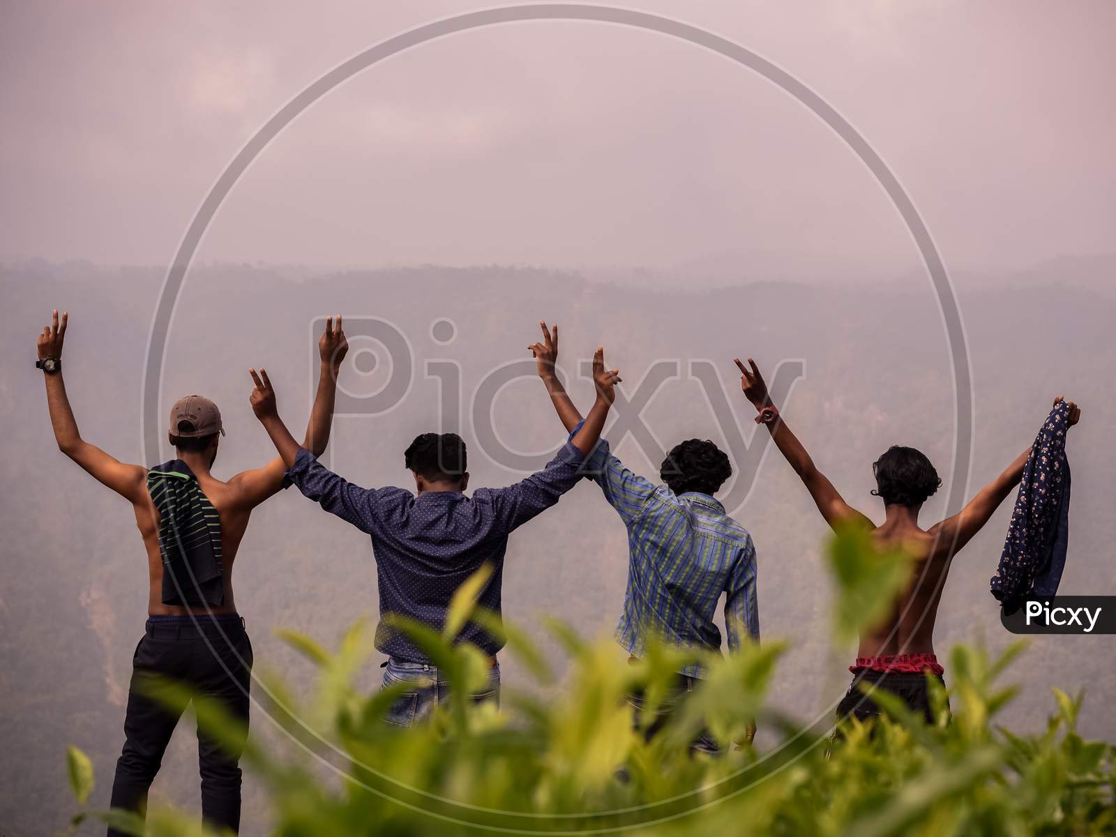 Silhouettes Of Group Of Four Happy Friends Is Enjoying And Having Fun At Mountain Top , Stands At Breath Taking View Point And Waving Victory Or Peace Sign And Raising Hands. Travel And Tourism.