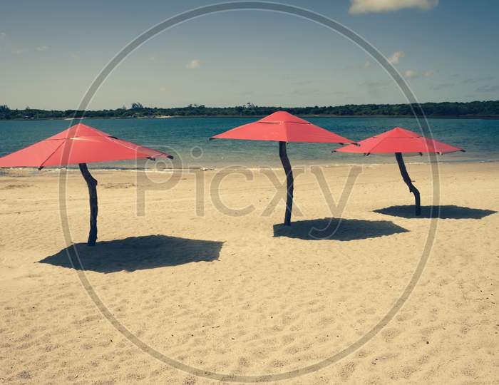 Red Umbrellas On The Beach Awaiting Tourists During A Sunny Day. Holiday Postcard.