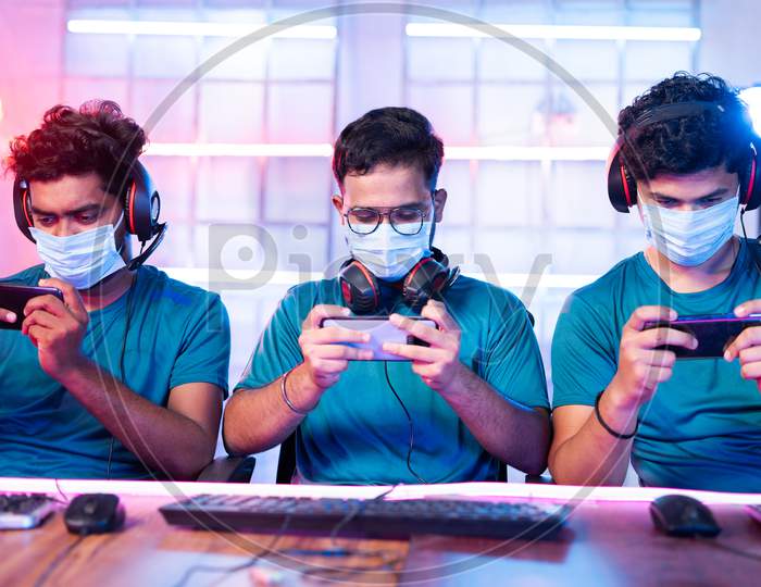 Group Of Gamers With Medical Face Mask Playing Video Game On Mobile Phone At Esports League Or Tournament During Coronavirus Or Covid-19 With Safety Measures.